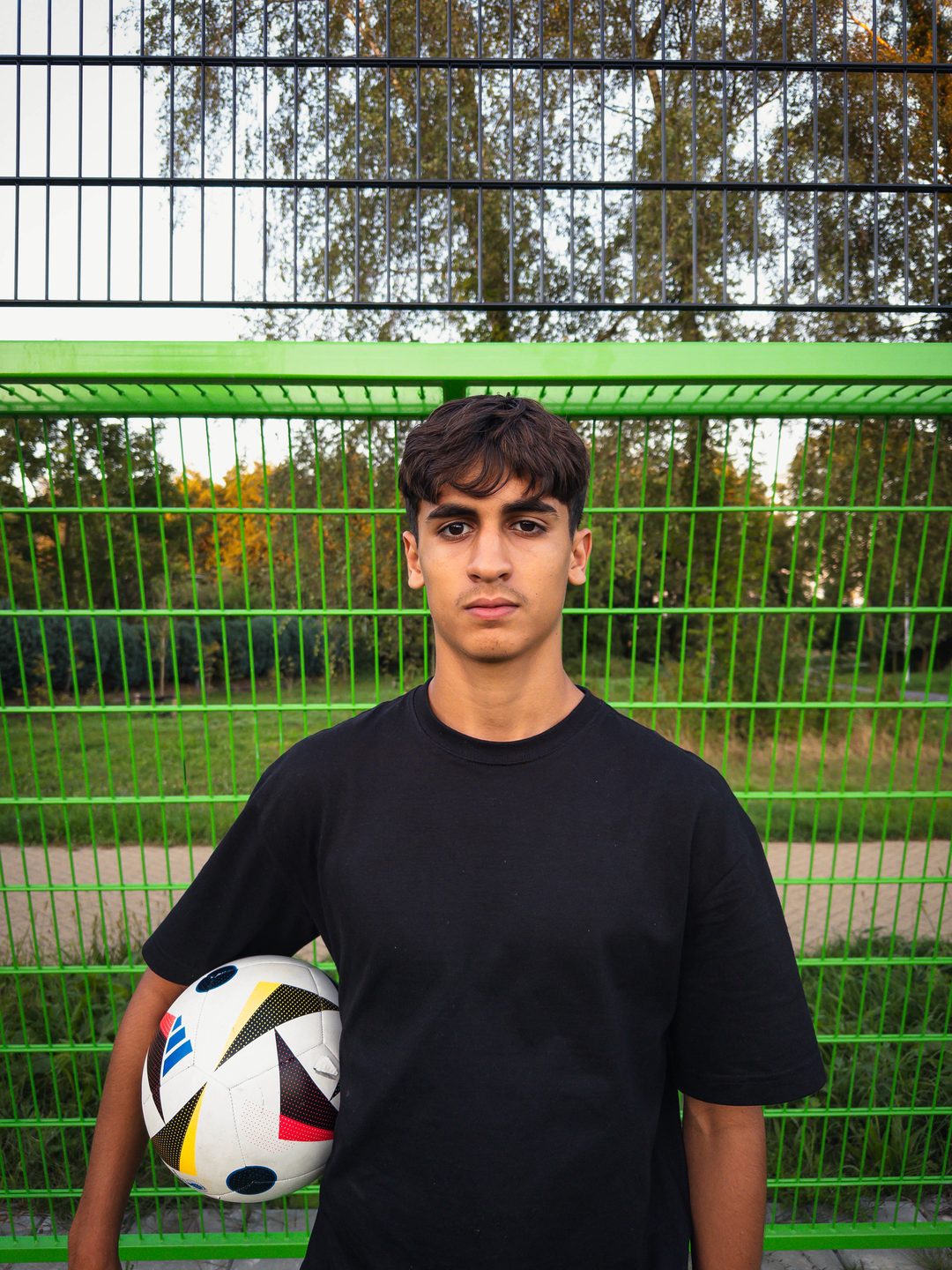 Portret van een jonge voetballer met bal voor een groen hek op een trapveld